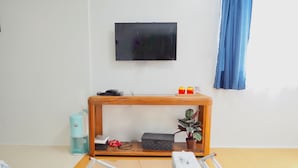 A living room with a wooden console table, a television mounted on the wall, a glass shelf with a plant, and a glass rack with magazines and a remote control. - Holiday Homes TSUBOYA 2nd (Naha)