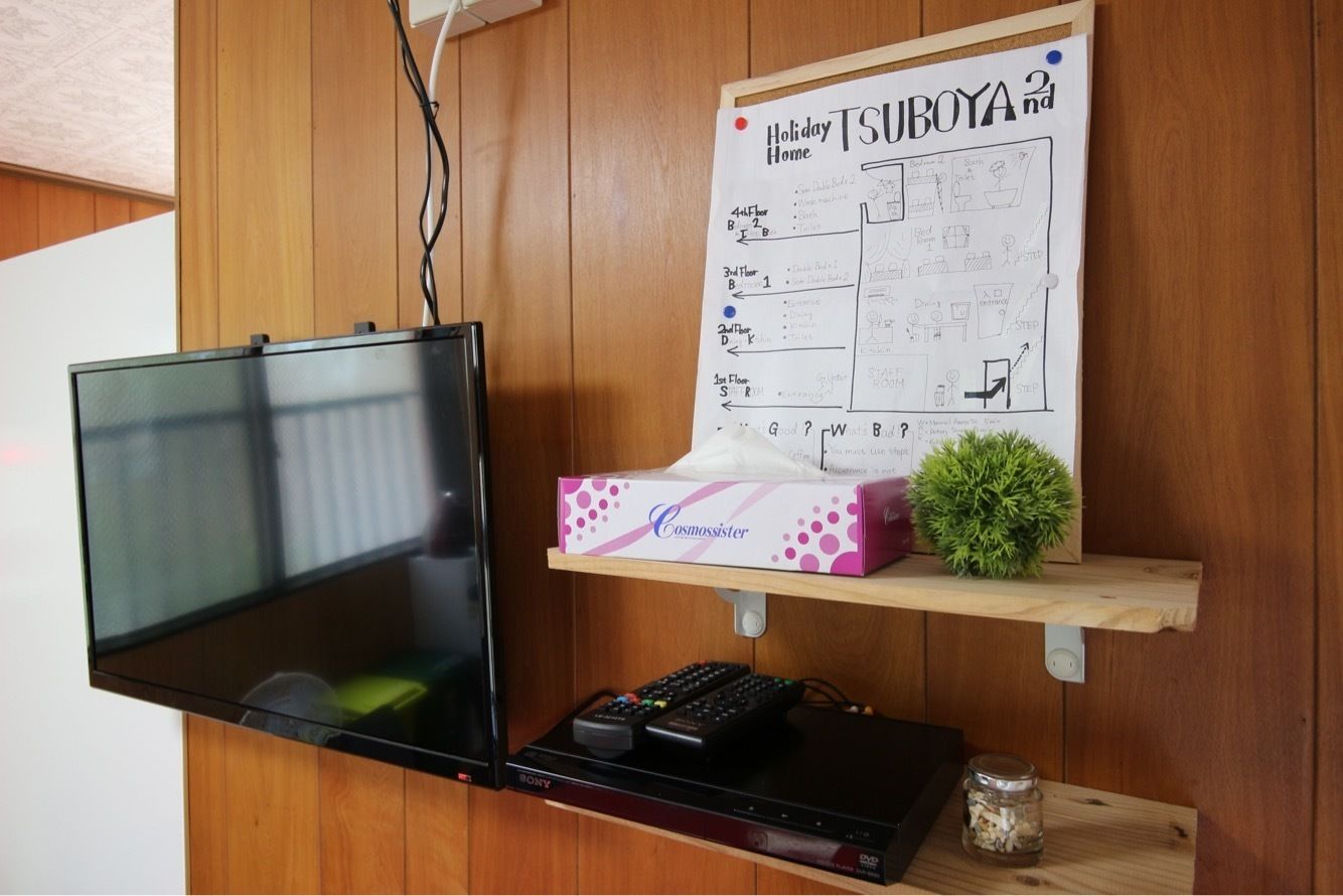 A flat-screen TV mounted on a wooden wall, a remote control, a DVD player, and a handwritten holiday home plan on a shelf.