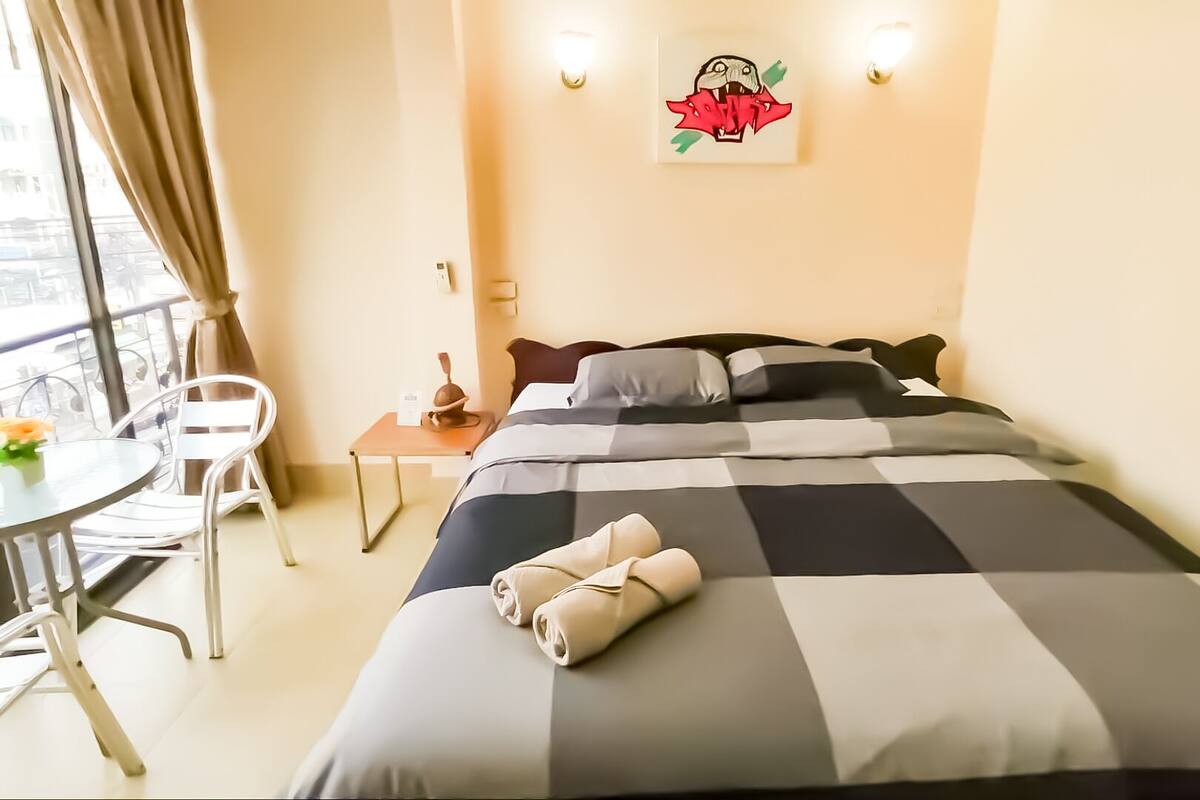 A hotel room with a bed, a small table with a chair, a window with curtains, and a wall-mounted light fixture.