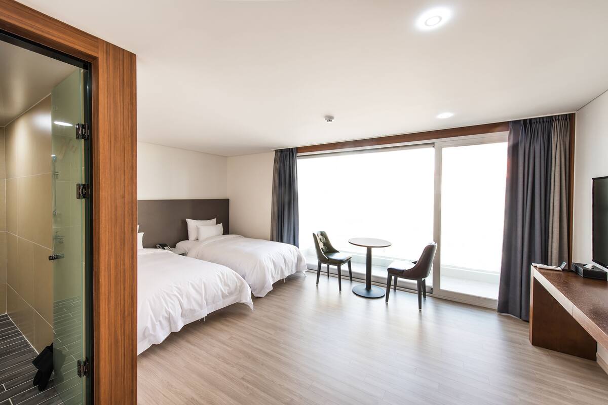 A hotel room with two beds, a glass shower enclosure, a dining area with a table and chairs, and a large window with curtains.