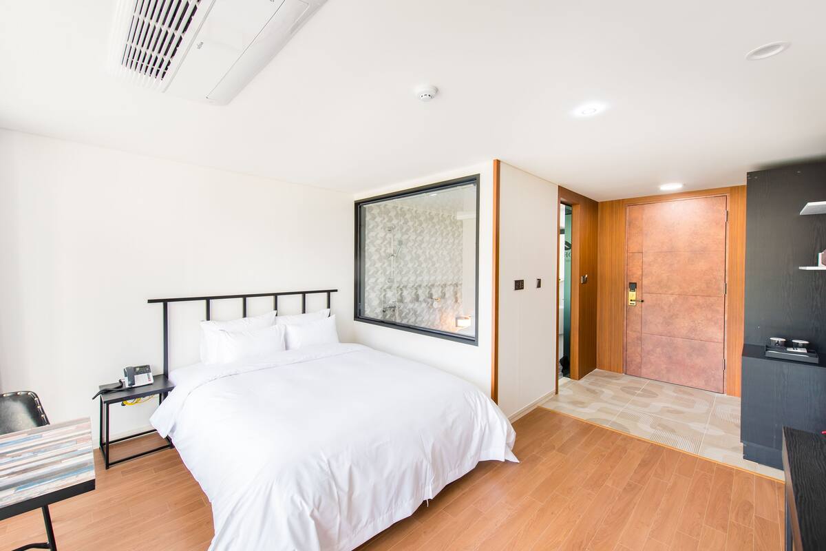 A modern hotel room with a large bed, a bedside table, a small desk, and a large window with a view of the outside.