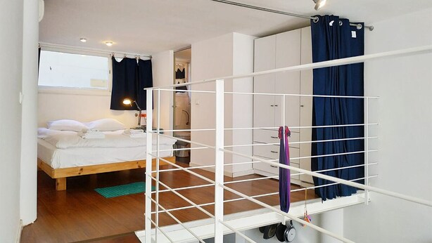 A bedroom with a bed, a white wardrobe, a drying rack, and a window with curtains. - AirTLV Centrally Located Studio With Gallery (Tel Aviv)