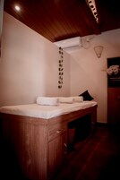 Un mueble de madera con cajones, una cama blanca con un colchón blanco y una toalla blanca enrollada, un aire acondicionado instalado en la pared y una lámpara fijada a la pared.