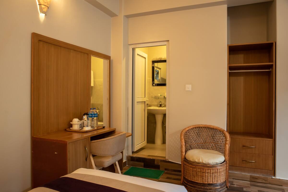 A hotel room with a wooden desk, a chair, a bed, a green mat, a wicker chair, and a wardrobe.