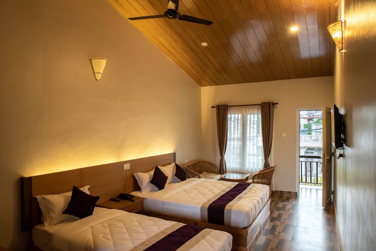 A hotel room with two beds, a ceiling fan, a small dining table, and a balcony with a view.