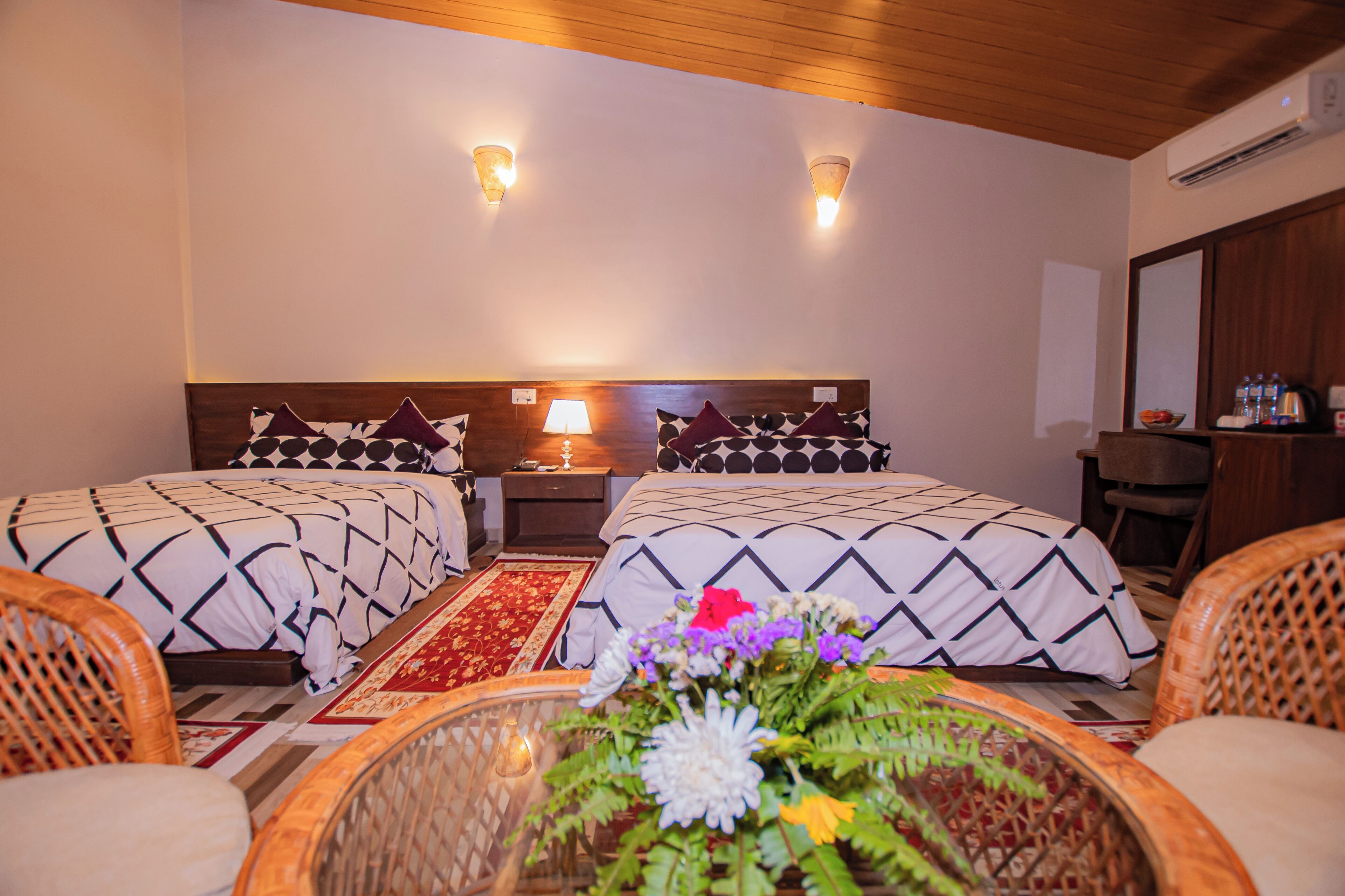 A hotel room with two beds, a wooden ceiling, a red and white patterned rug, wicker chairs, and a table with a floral arrangement.