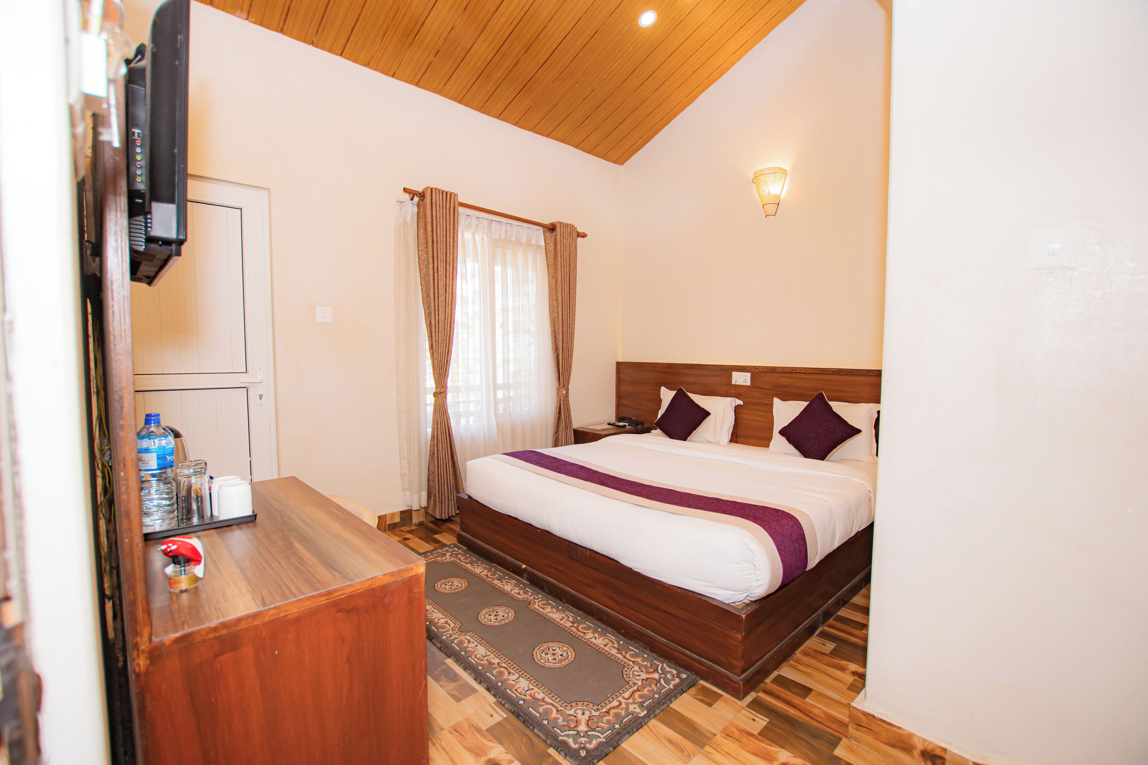 A hotel room with a wooden ceiling, a bed with white and purple bedding, a bedside table with a water bottle, and a small rug.