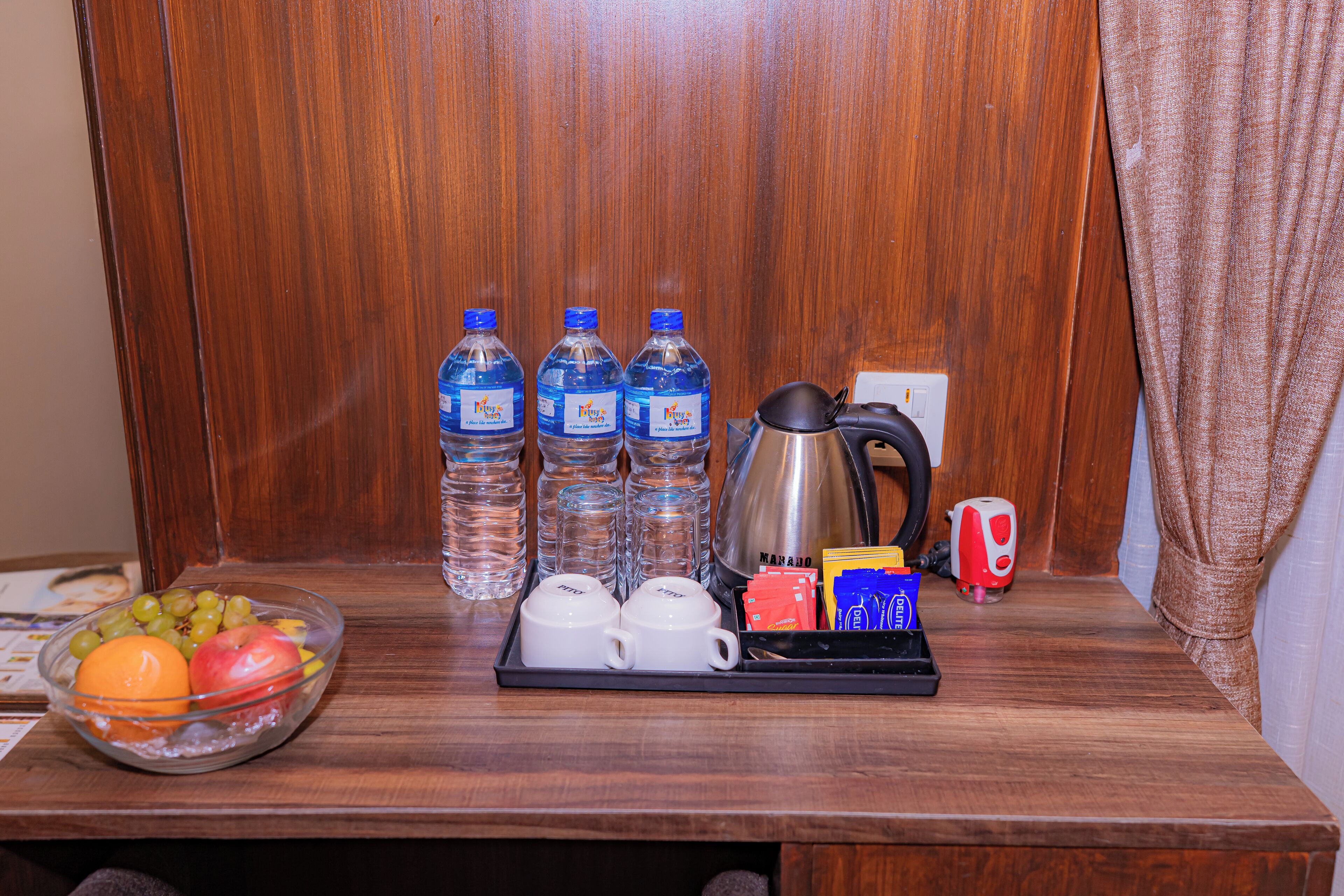 A wooden table with a fruit bowl, water bottles, coffee cups, a kettle, and a tray with coffee packets and a mug.