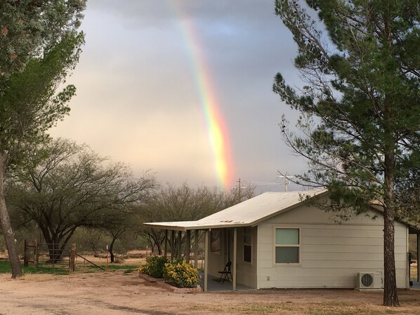 A single-story building with a porch, a small garden, and a visible rainbow in the background.