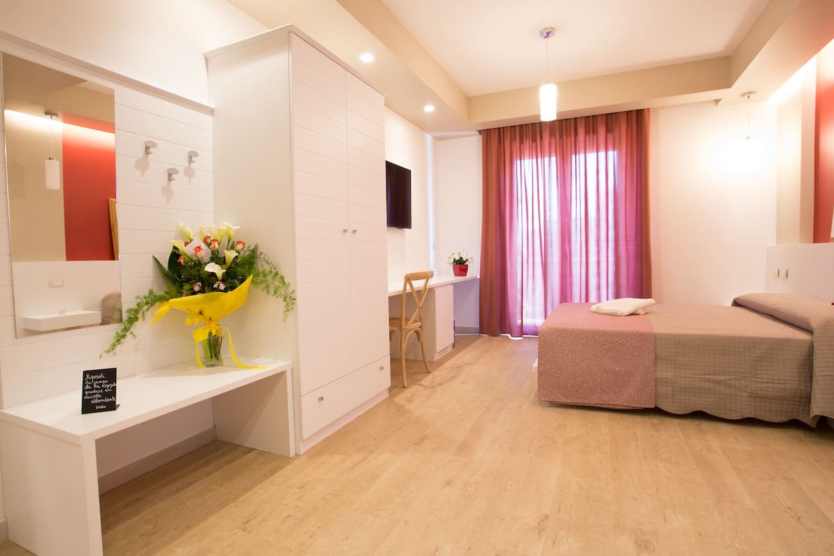 Una habitación de hotel moderna con piso de madera, una cama con cubrecama rosa, un escritorio con silla, una mesita con planta en maceta y un espejo grande en la pared.
