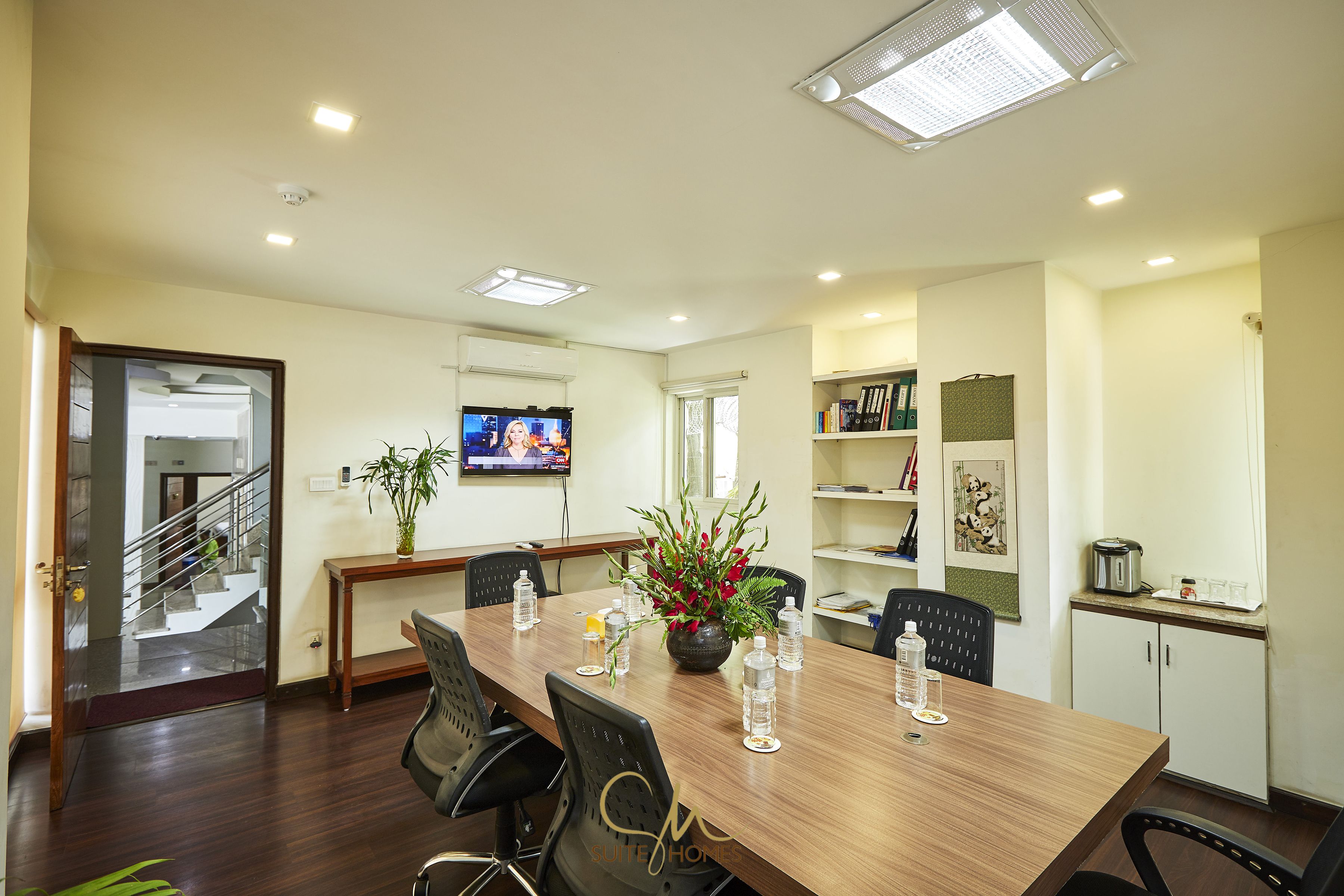 Un salón de conferencias con una mesa de madera, sillas, un televisor, una planta y botellas de agua.