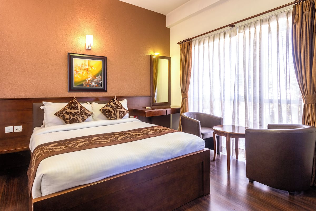 Habitación de hotel con una cama grande, un escritorio, una silla y vistas de la ciudad a través de cortinas translúcidas.