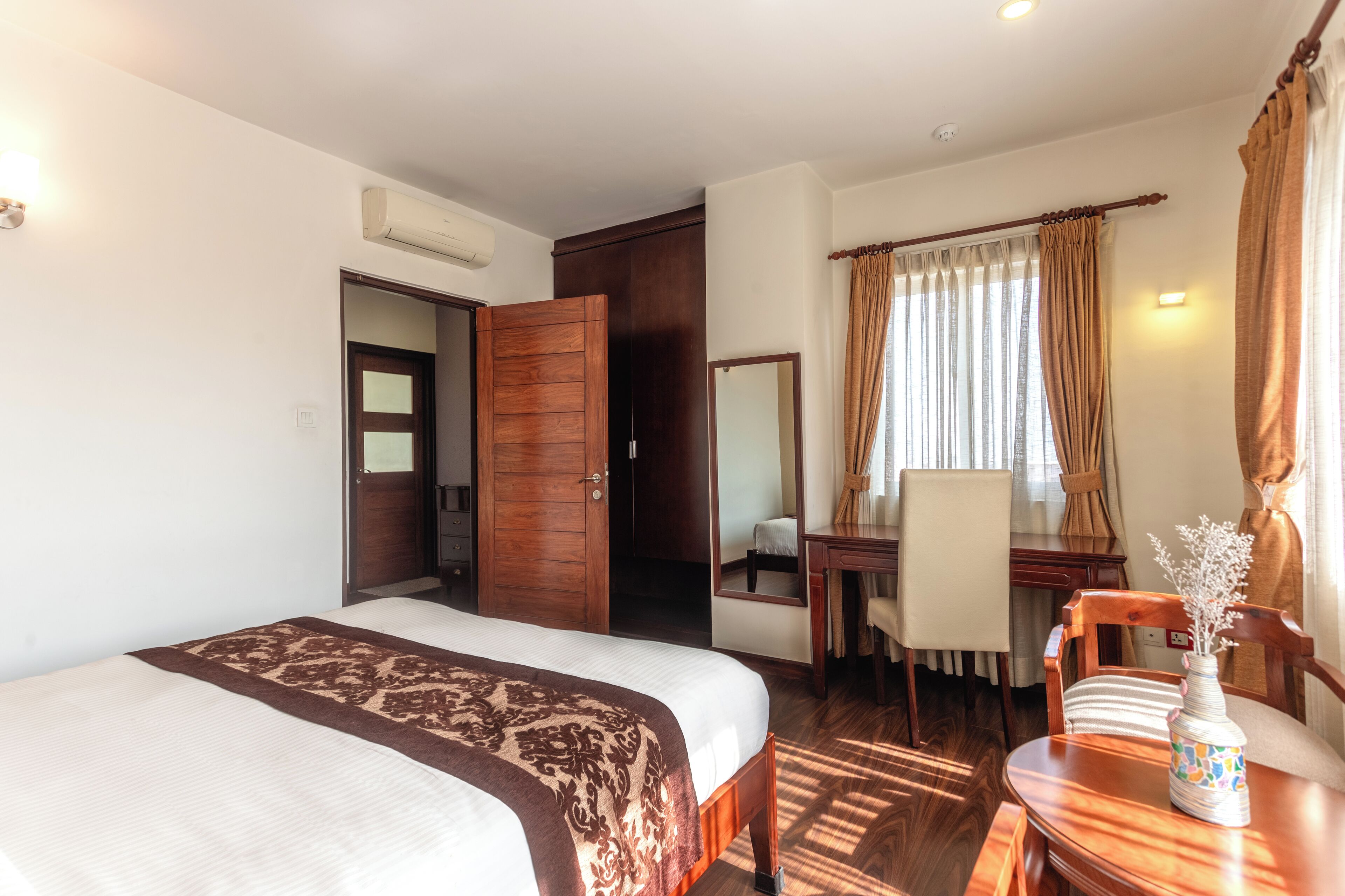 Une chambre d’hôtel avec un lit, un bureau, une chaise, un miroir et une table sur laquelle se trouve un vase.