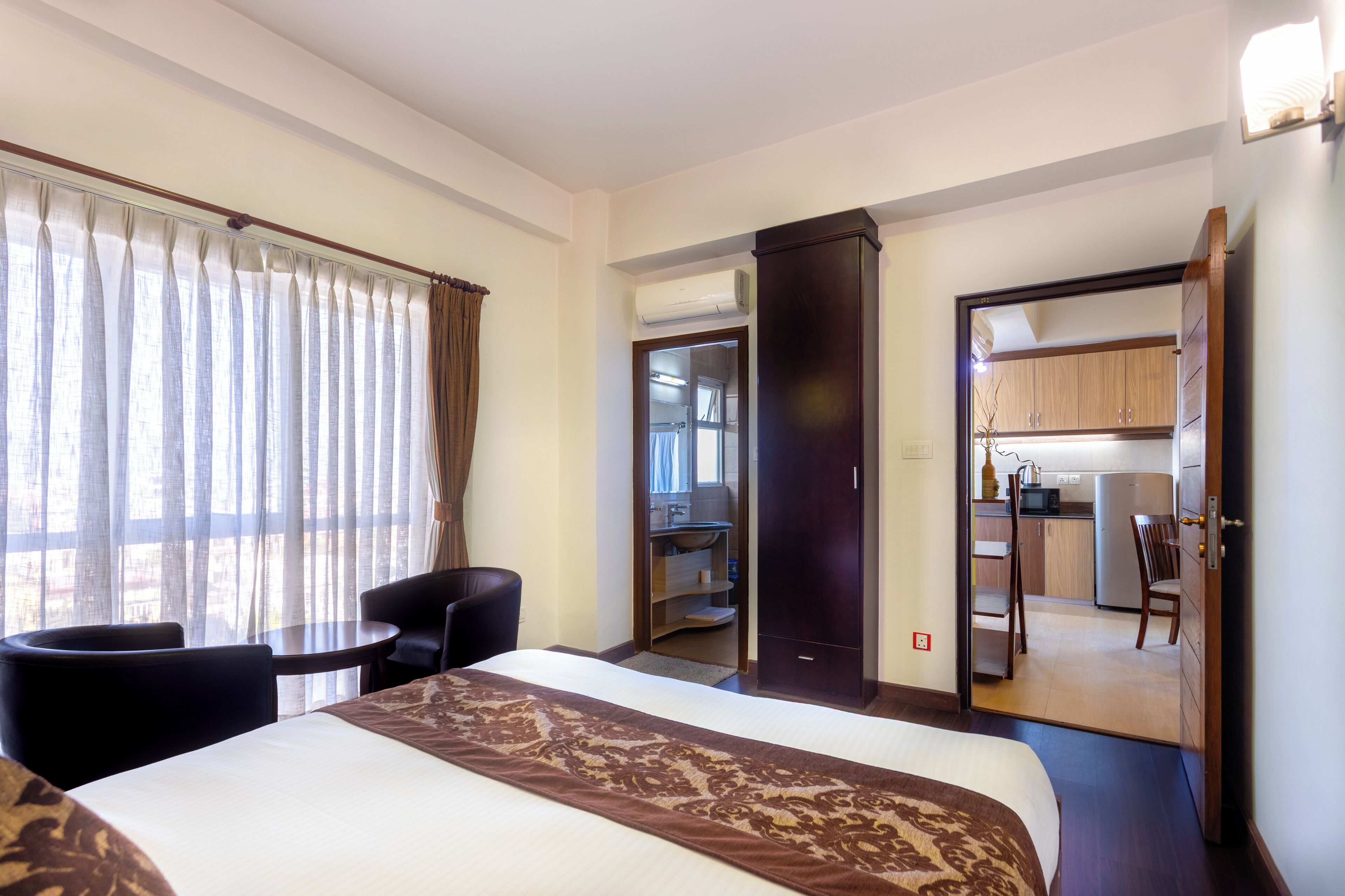 A hotel room with a bed, a small table, a chair, a kitchenette, and a bathroom visible through an open door.
