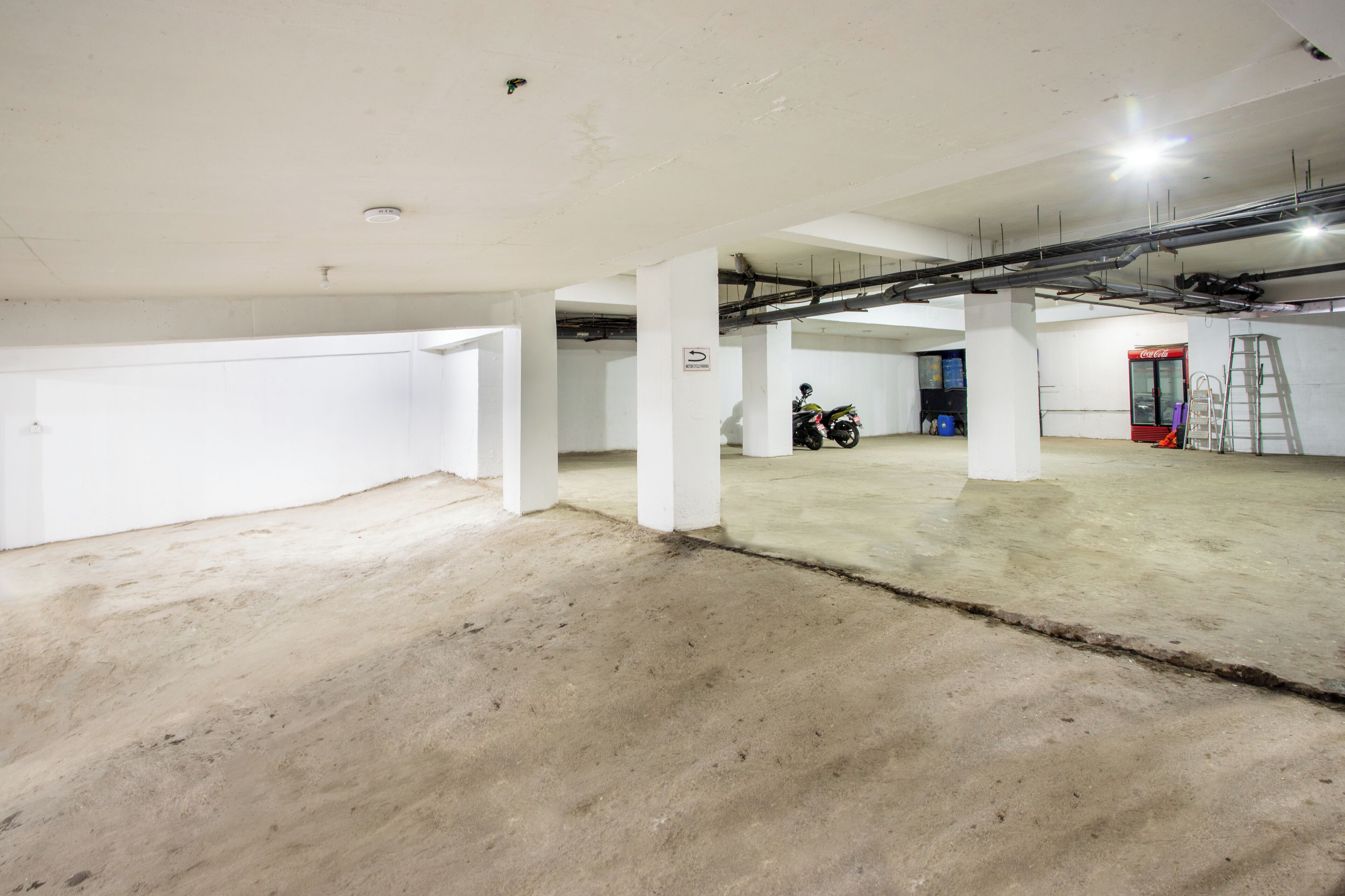 A spacious indoor parking area with concrete floors and white walls, featuring a motorcycle and a red Coca-Cola refrigerator.
