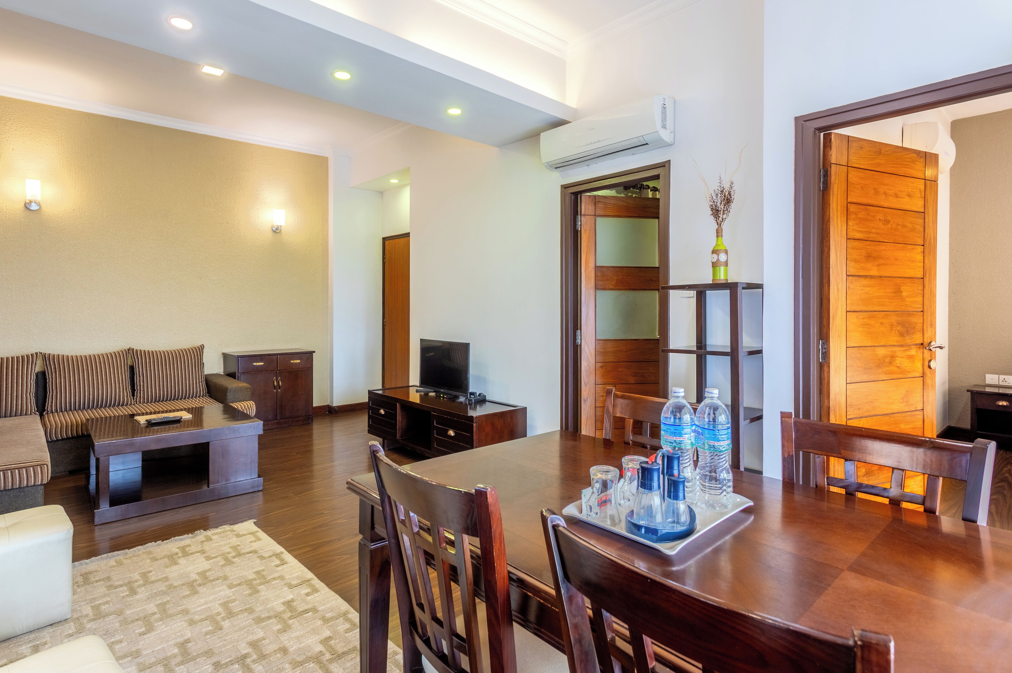 Una sala de estar amplia con sofá, una mesa de centro y televisor, un comedor con mesa y sillas de madera, y una puerta de madera con un banco.