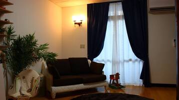 A living room with a dark brown sofa, a small wooden chair, a large potted plant, a small table, and a window with dark curtains.