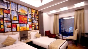 A hotel room with two beds, a large abstract painting on the wall, a red chair, a white armchair, and a view of the outdoors through a window. - Hotel City Pride (New Delhi)