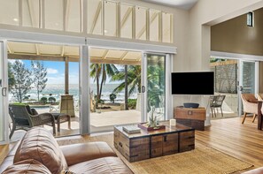 A spacious living room with a large sliding glass door leading to a balcony with a view of the ocean. There is a comfortable seating area with a sofa and chairs, a coffee table, and a flat-screen TV. - Oceans Edge Beach House At Oak Beach (Oak Beach)