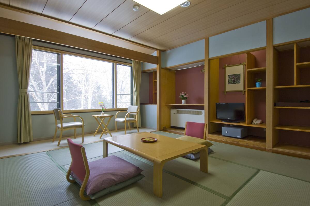 Una habitación con esteras de tatami, una mesa de madera, sillas, un televisor y un estante con objetos.