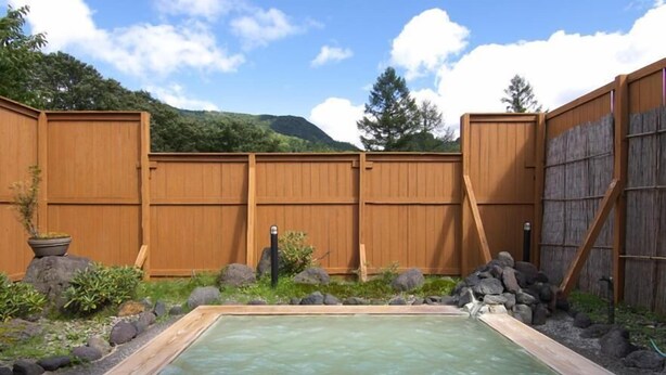 A rectangular hot tub with a wooden frame, surrounded by a wooden fence, rocks, and greenery. - Hotel Hananoki (Nikko)