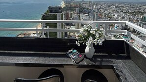 Ein Balkon mit Blick auf das Meer und die Stadtlandschaft, zwei Barhocker und eine Vase mit Blumen.
