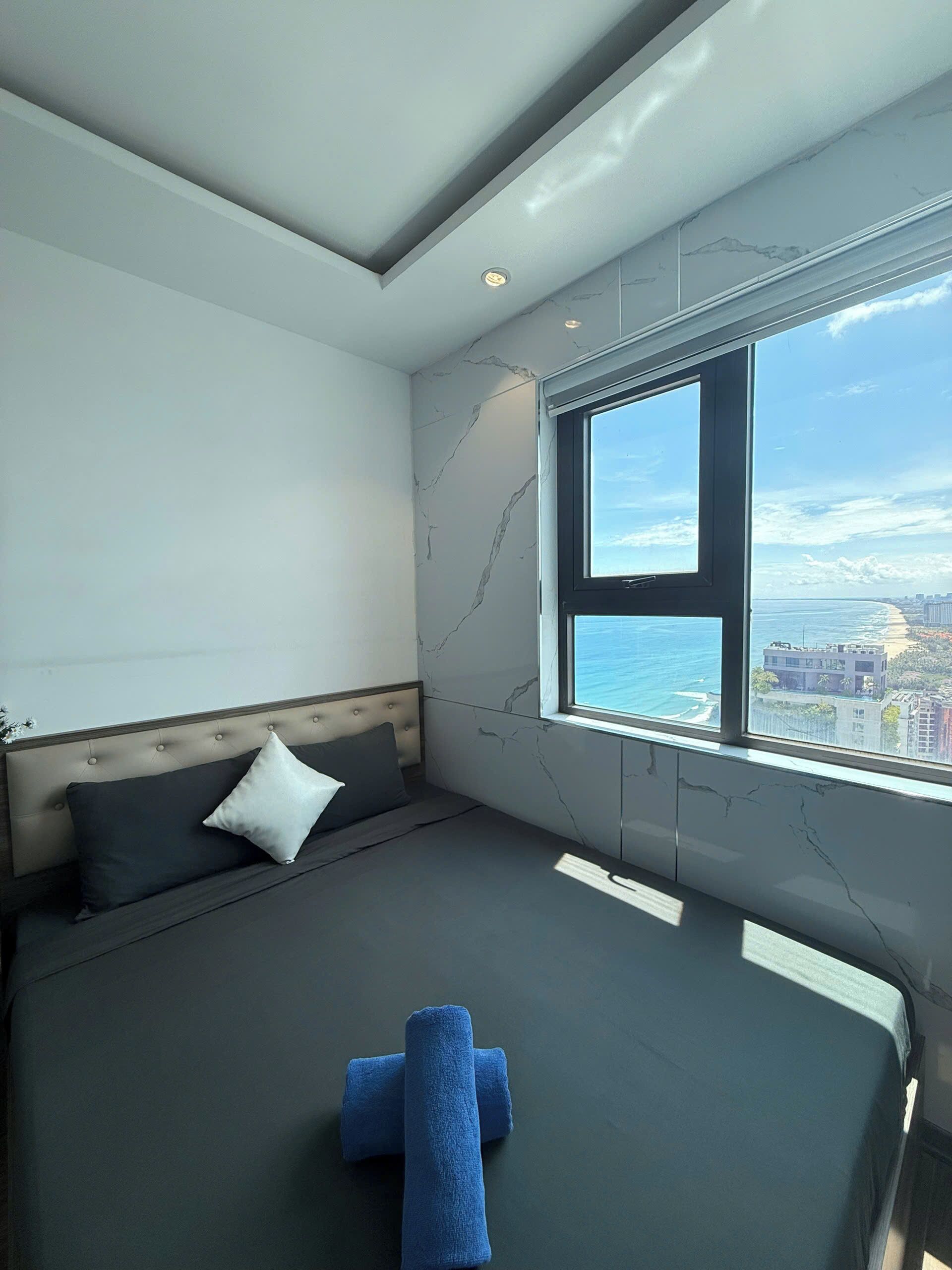 A modern bedroom with a large window, a bed with dark bedding, and a view of the sea and cityscape.