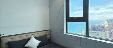 A modern bedroom with a large window, a bed with dark bedding, and a view of the sea and cityscape.