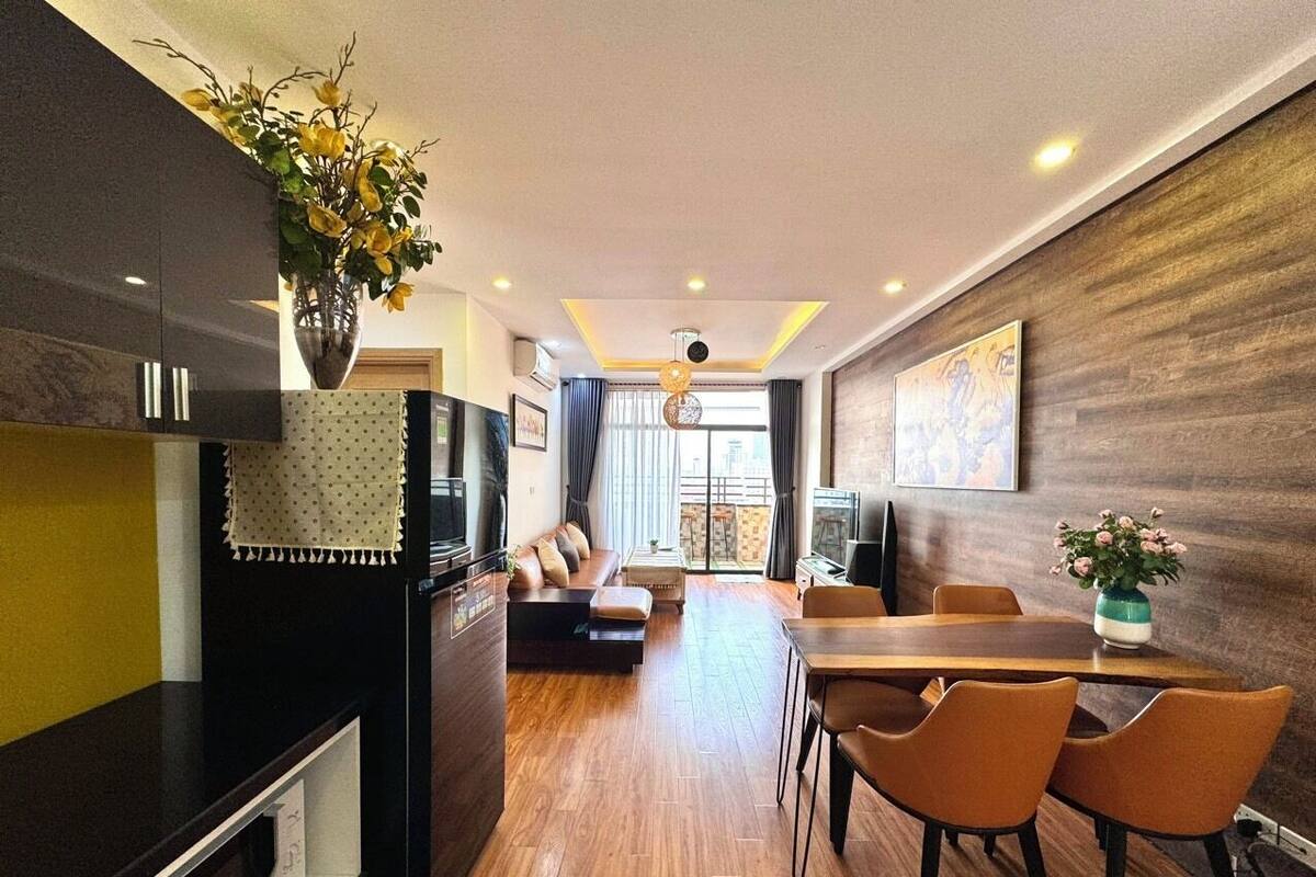 A modern living room with a sofa, dining table, and chairs. There is a kitchen area with a refrigerator and a vase of flowers.