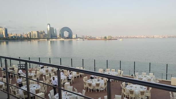 Ein Restaurant am Wasser mit Tischen und Stühlen, das auf ein Gewässer und eine Stadtkulisse im Hintergrund blickt.