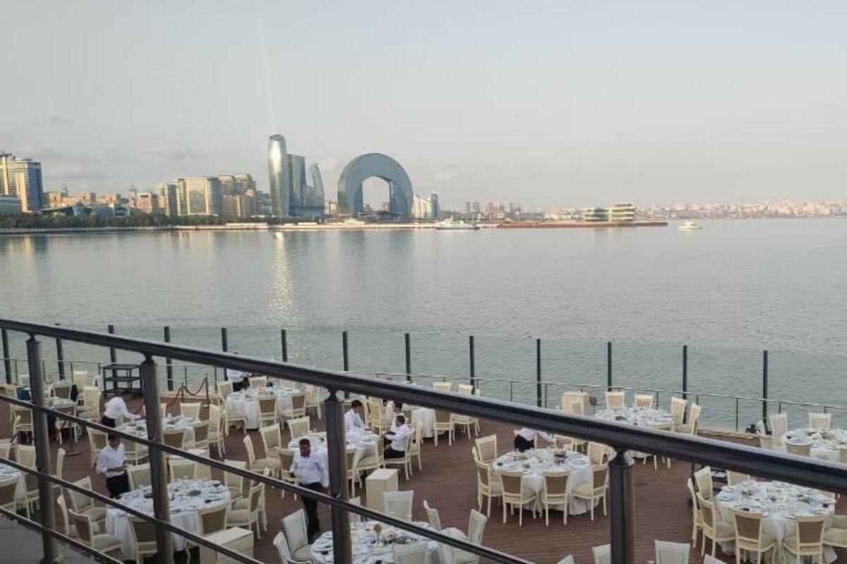 Un restaurante frente al agua, con mesas y sillas dispuestas para comer, con vista a un cuerpo de agua y un horizonte urbano al fondo.
