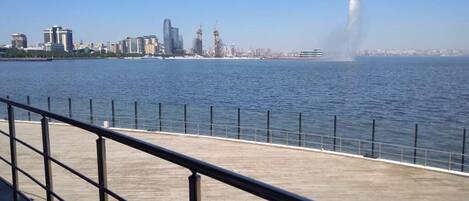 A waterfront promenade with a metal railing, a city skyline in the background, and a fountain in the distance.