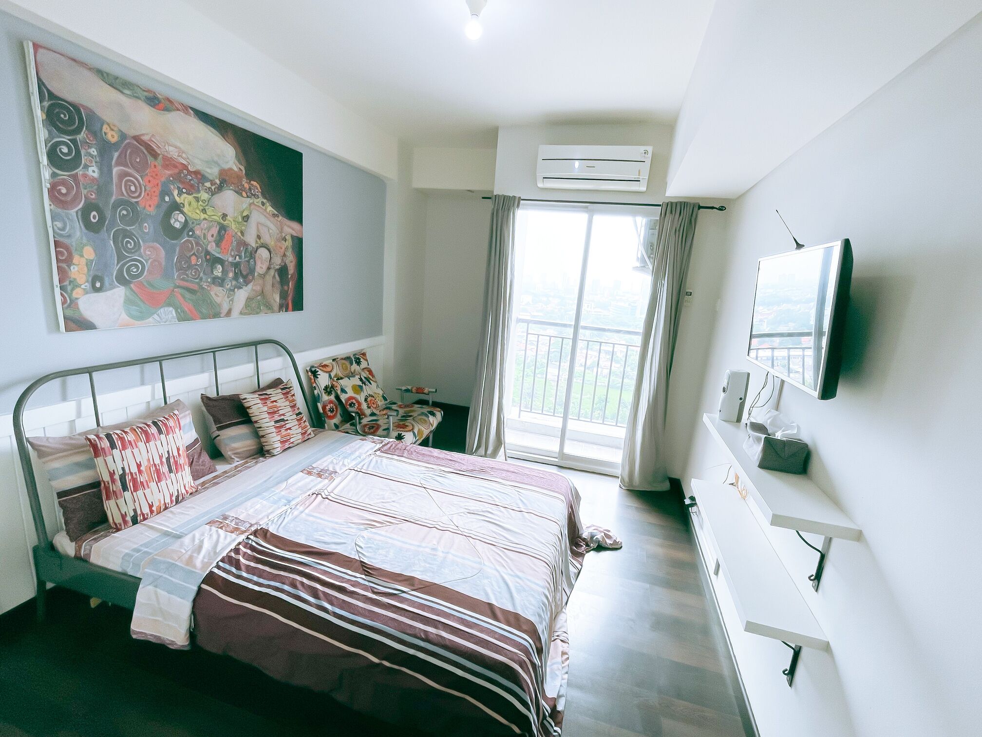 A bedroom with a bed, a painting, an air conditioner, a TV, and a balcony with curtains.