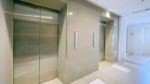 A modern elevator with a metallic finish, adjacent to a wall with a light switch and a door marked with the number 17-12. - The Satu Stay Apartement SGV (South Tangerang)