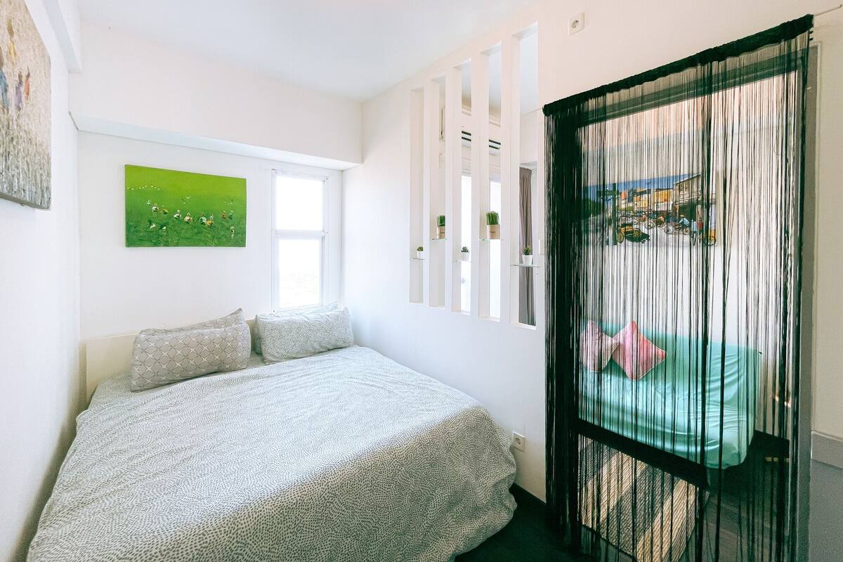 A bedroom with a bed, a window, and a view of a cityscape through a black curtain.