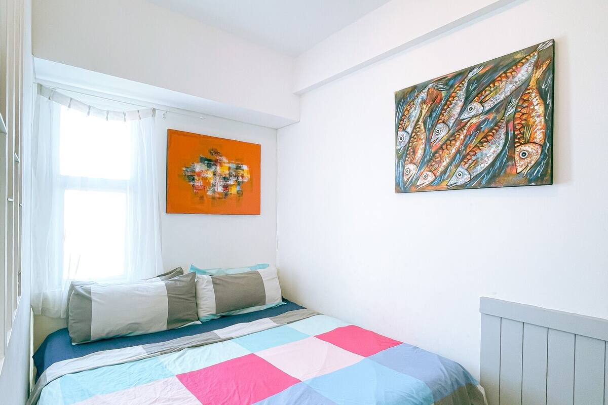 A bedroom with a bed, a window with curtains, a radiator, and two paintings on the wall.
