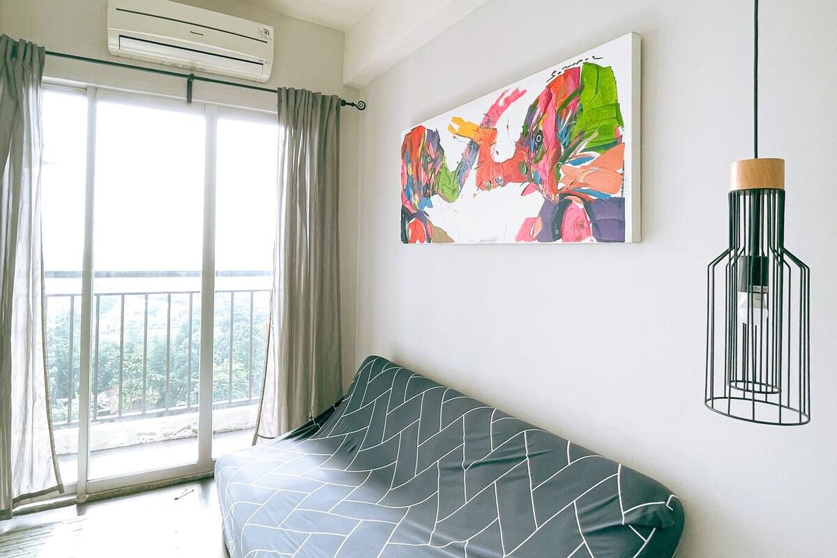 A modern living room with a sofa, a large window with curtains, an air conditioning unit, a colorful abstract painting, and a pendant light.