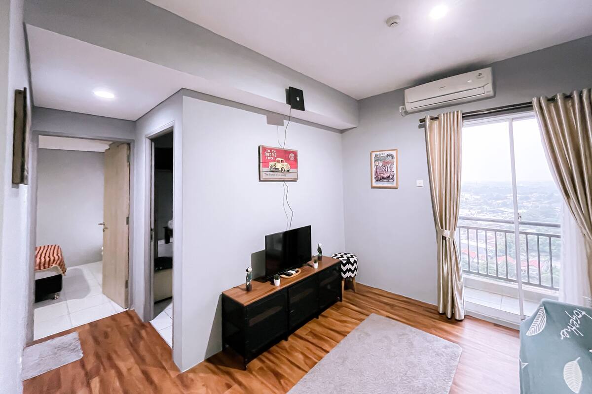 A modern living room with a flat-screen TV, wooden flooring, a sofa, and a large window with curtains.
