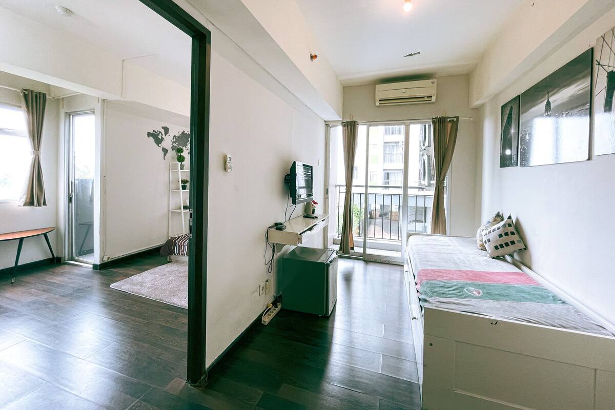 A modern hotel room with a bed, a desk, a chair, a small table, a plant, and a window with curtains.