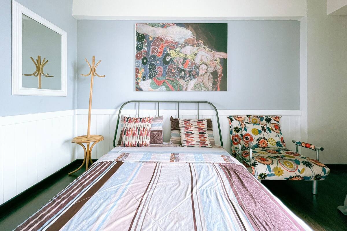 A bedroom with a bed, a chair, a coat rack, and a painting on the wall.
