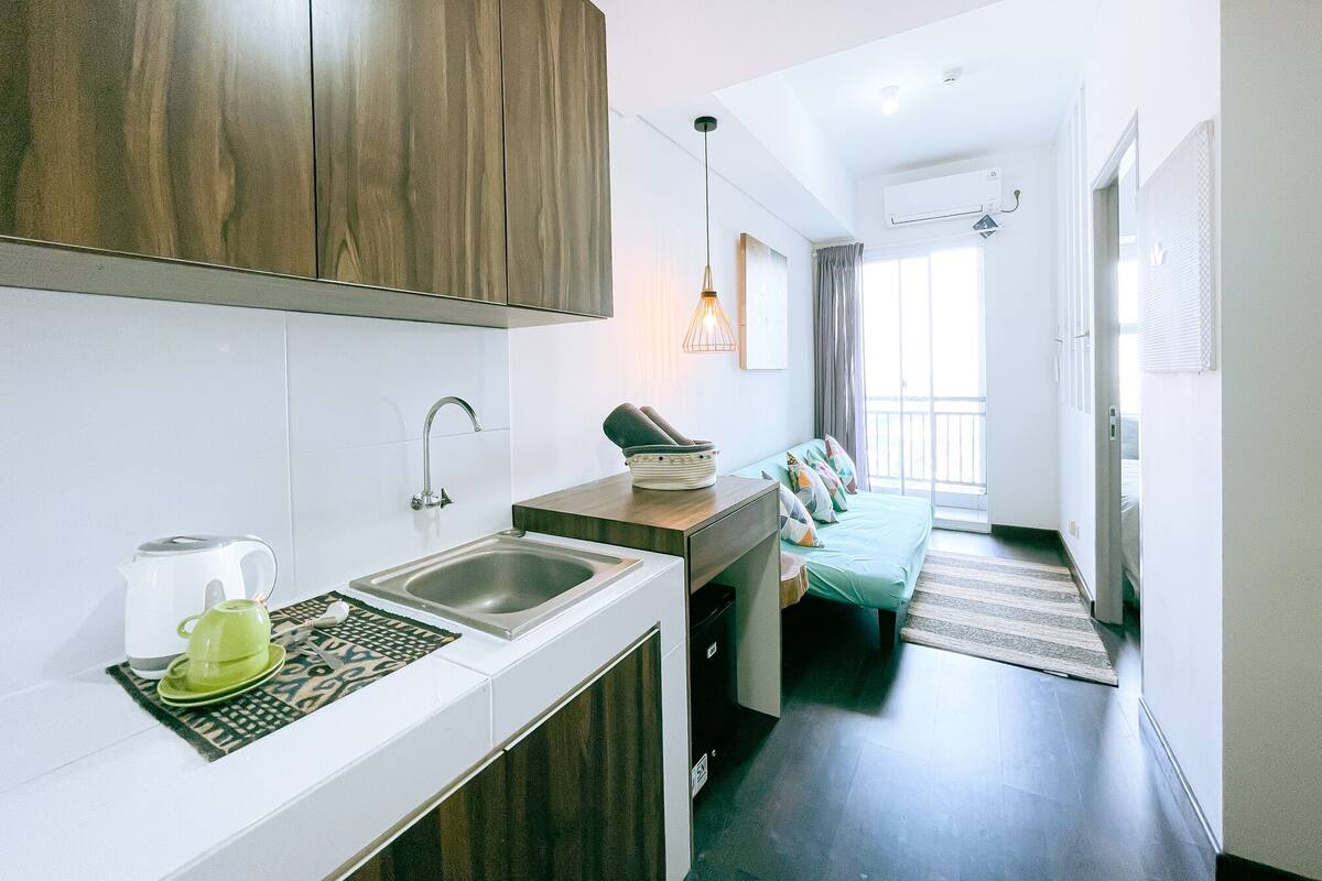 A modern kitchen with a sink, a kettle, and a coffee maker. There is a sofa with cushions in the adjacent living area.