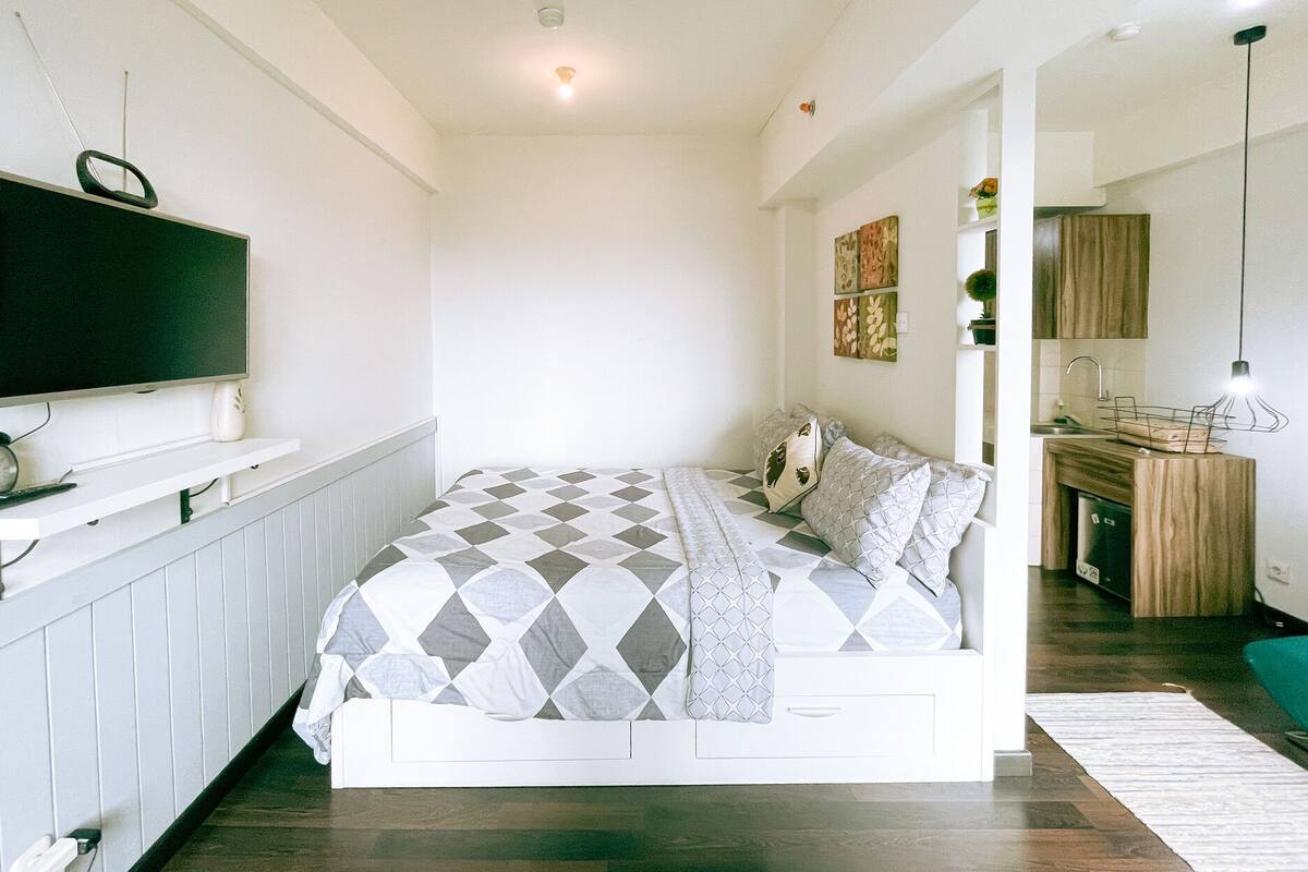 A bedroom with a bed, a TV, a bedside table, a mirror, and a kitchen area.