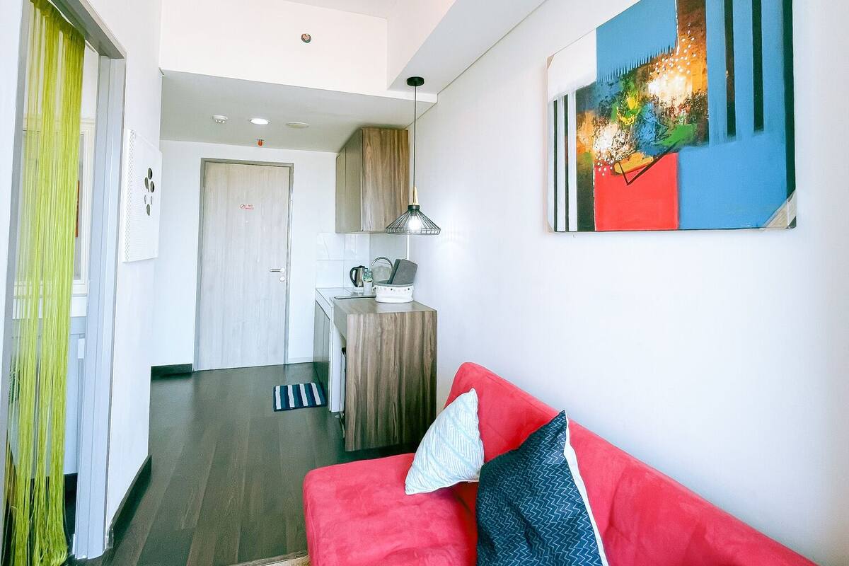 A modern living room with a red sofa, a kitchen area in the background, and a colorful abstract painting on the wall.