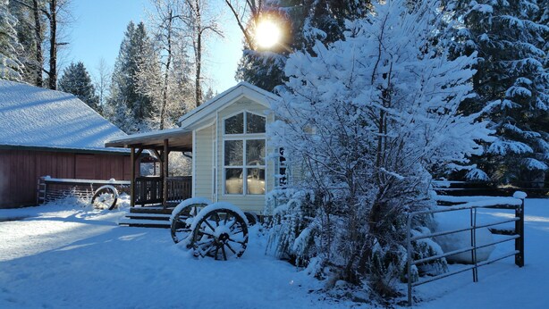 A snow-covered cabin with a wooden porch, a large wheel, and a fenced area.