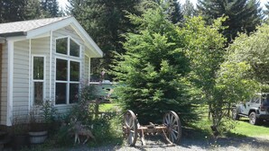A rustic cabin with a large window, surrounded by dense greenery and a wooden cart.