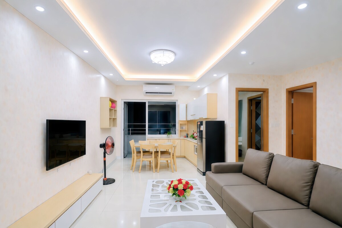 A modern living room with a flat-screen TV, a sofa, a dining table with chairs, a kitchen area, and a ceiling light.