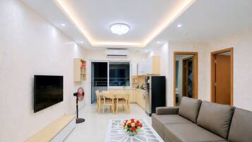 A modern living room with a flat-screen TV, a sofa, a dining table with chairs, a kitchen area, and a ceiling light.