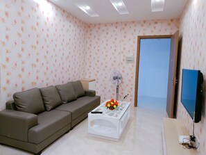 A living room with a sofa, a coffee table with a vase, a standing fan, and a TV on a stand. - HQH Sea View Apartment (Bac Nha Trang)