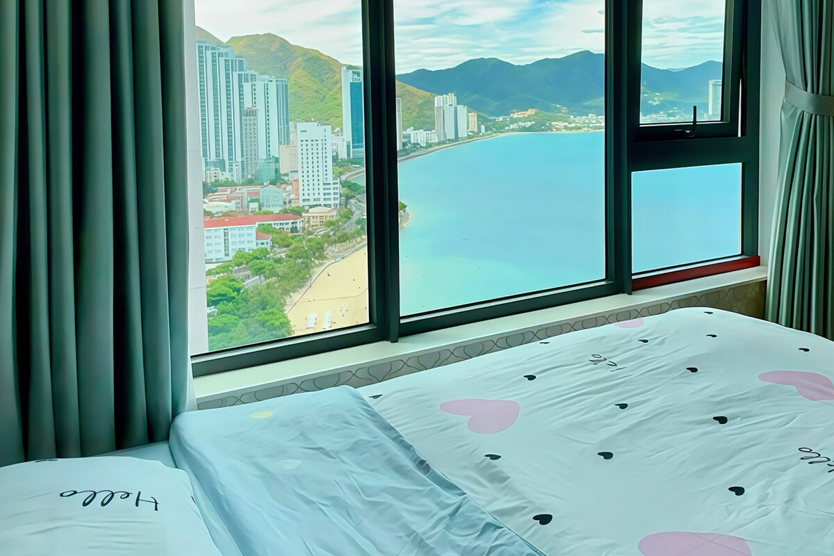 A hotel room with a large window offering a view of a coastal cityscape and mountains.