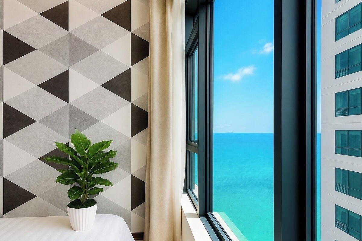 A hotel room with a bed, a potted plant, a window with a view of the sea, and a geometric-patterned wall.